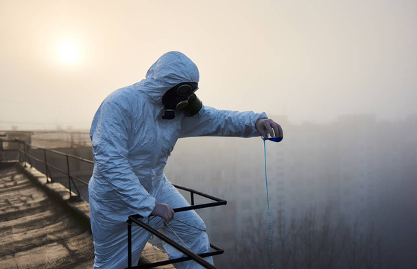 Wissenschaftler in Schutzkleidung und Gasmaske, arbeitet auf dem Dach eines Hochhauses, führt die Studie durch und verwendet blaues Reagenz in Glaskolben bei Sonnenaufgang - Foto, Bild