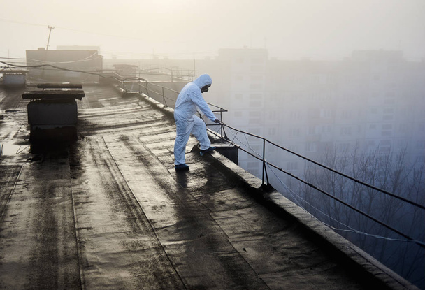 Researcher in protective outfit and gas mask, thinking of important environmental problems and their influence on people's lives, standing on the roof of high building in foggy morning - Foto, afbeelding