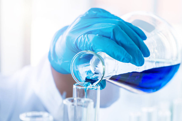 Main de scientifique avec un gant en caoutchouc versant du liquide chimique bleu de la fiole en verre pour tester le tube. Science concept biochimique et pharmaceutique
. - Photo, image