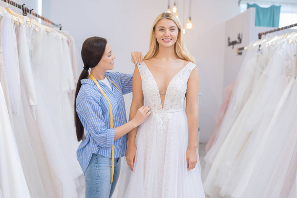 Lachende trouwjurk kleermaker met tape maatregel aanpassen jurk gemonteerd door jonge bruid in bruiloft showroom - Foto, afbeelding
