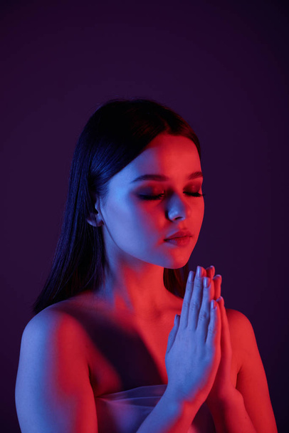 Serene calm young woman with naked shoulders meditating with closed eyes against dark blue background, neon light - Fotó, kép