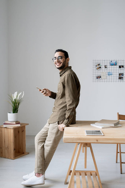 Jovem empresário alegre usando smartphone enquanto está à mesa na frente da câmera no escritório
 - Foto, Imagem