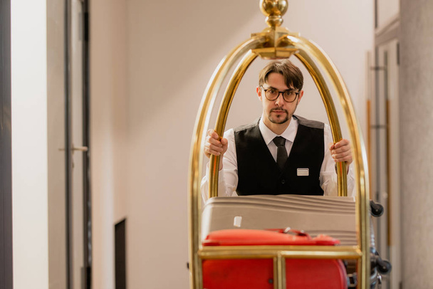 Jovem porteiro elegante em óculos empurrando carrinho com bagagem de hóspedes do hotel, enquanto se move para a frente ao longo do corredor - Foto, Imagem