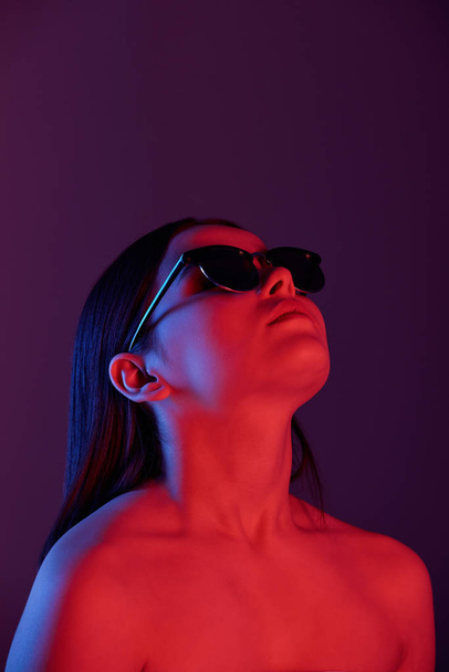 Vakava alaston nuori nainen aurinkolaseissa katselee tumman neonvalon alla
 - Valokuva, kuva