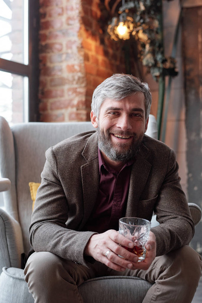 Portrait d'un bel homme positif en veste brune assis dans un fauteuil et dégustant du whisky à la maison
 - Photo, image