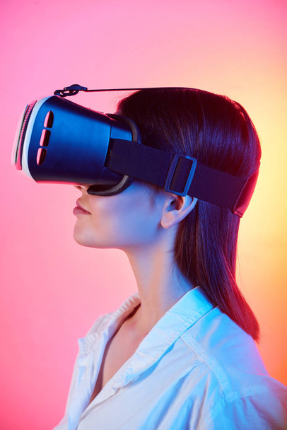 Zijaanzicht van brunette meisje in witte blouse met behulp van virtual reality bril tegen tender roze en gele achtergrond - Foto, afbeelding