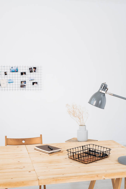 Mesa de madeira com lâmpada, planta em vaso, notebook com caneta e outros suprimentos contra parede branca no escritório
 - Foto, Imagem