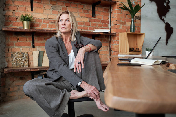 Barefoot mature businesswoman with blond hair sitting in relaxed pose on chair in office and being deep in thoughts - Photo, Image