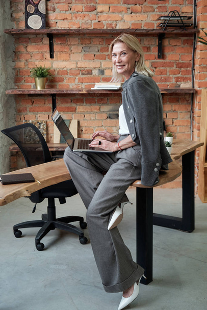 Πορτρέτο του χαρούμενα επιτυχημένη ώριμη επιχειρηματίας κάθεται στο τραπέζι και χρησιμοποιώντας σύγχρονο φορητό υπολογιστή στο γραφείο - Φωτογραφία, εικόνα