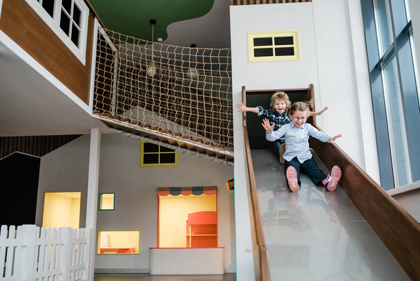 Zwei niedliche fröhliche Kinder in Freizeitkleidung bewegen sich die Rutsche hinunter, während sie sich auf dem Spielplatz im Kinderzentrum vergnügen - Foto, Bild