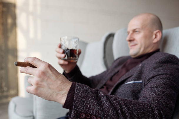 Şık ceketli, rahat kel bir adamın yakın çekimi koltukta oturup viski içerken puro içmesi. - Fotoğraf, Görsel