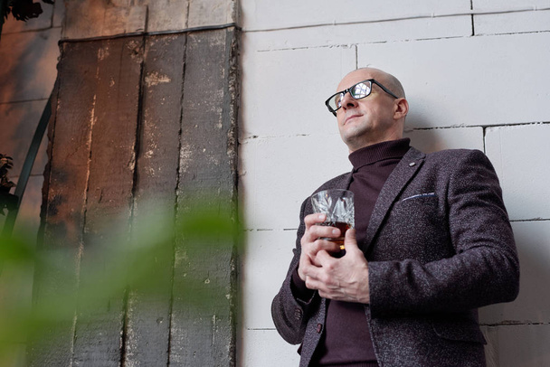 Below view of sad contemplative bald man in glasses standing at wall and drinking alcohol - Photo, Image