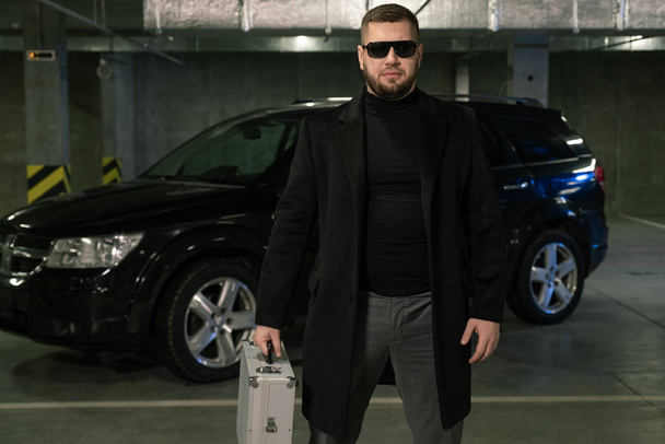 Bärtiger Krimineller oder Agent in schwarzem Mantel und Sonnenbrille bewegt sich mit Koffer mit Bargeld oder Drogen über den Parkplatz - Foto, Bild