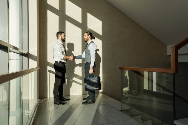 Two young elegant business partners handshaking after negotiating and signing new contract while standing against wall - 写真・画像
