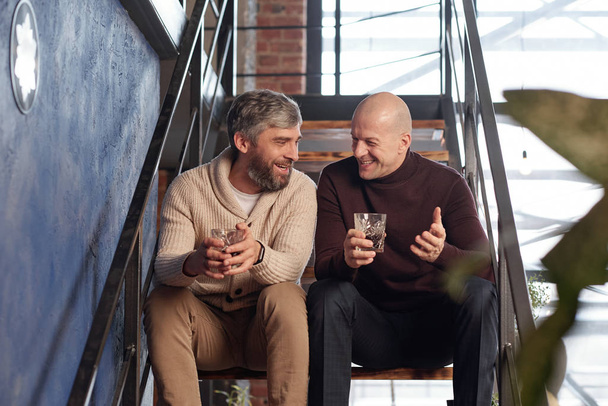 Positive middle-aged men in casual outfits sitting on staircase and drinking alcohol while having fun together - Photo, Image