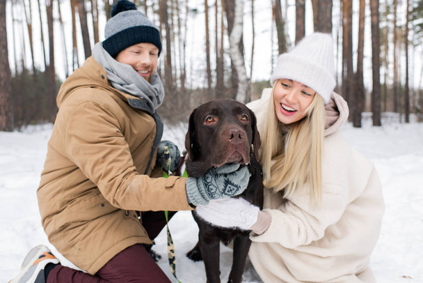 Happy young affectionate couple playing with black retriever sitting on snow while enjoying winter day in park or forest - Photo, image