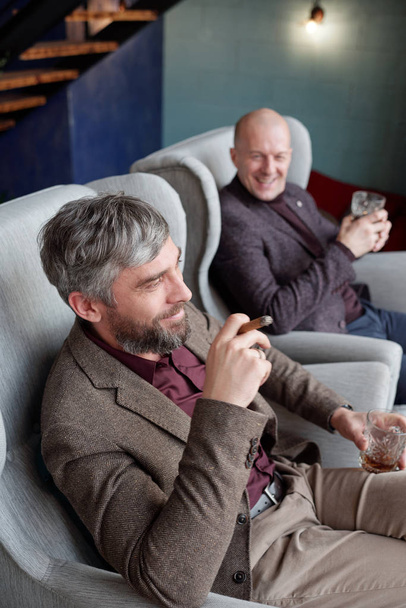 Positive rich middle-aged men in jackets sitting in armchairs and smoking cigars in living room - Фото, изображение