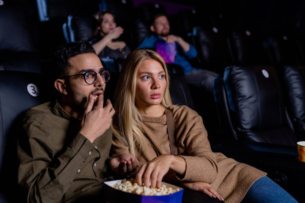 Young scared dates with box of popcorn sitting in dark cinema and watching horror movie at leisure - Valokuva, kuva