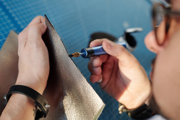 Nahaufnahme eines Handwerkers, der während der Verarbeitung von Leder in der Werkstatt schwarze Farbe auf den Lederrand aufträgt - Foto, Bild