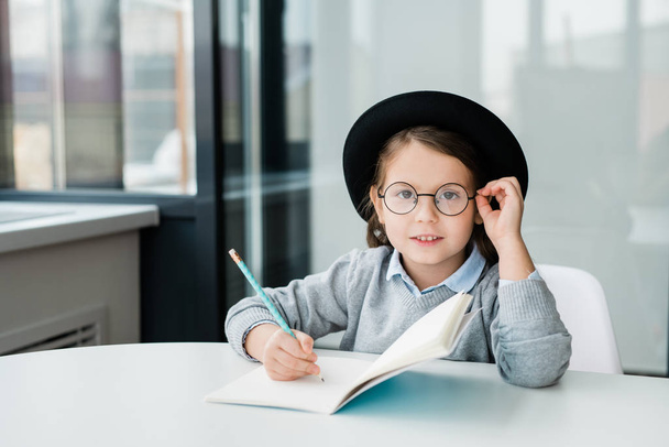 Schattig slim schoolmeisje met hoed die door een bril naar je kijkt terwijl ze aan het bureau zit en aantekeningen maakt in het copybook. - Foto, afbeelding