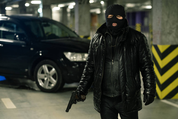 Terrorismo o gángster con chaqueta negra, guantes y balaclava portando pistola con coche esperando en segundo plano. - Foto, Imagen