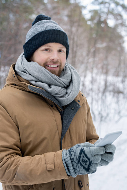 Young man in grey beanie, scarf, gloves warm jacket looking at you while scrolling in smartphone on winter day - Fotoğraf, Görsel