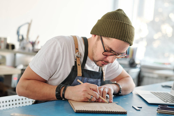 Poważny młody człowiek na rysunkach okularów w szkicowniku podczas tworzenia projektu produktu skórzanego w warsztacie - Zdjęcie, obraz