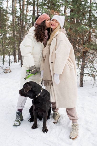 Happy girl kissing another one on cheek while both enjoying walk with black retriever on winter day in snowbound park - Foto, Imagen