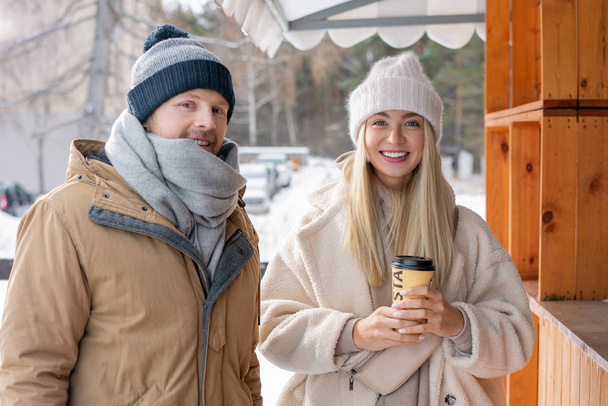 Happy young heterosexual couple in casual winterwear standing by wooden stall and looking at you while chilling out - Photo, Image