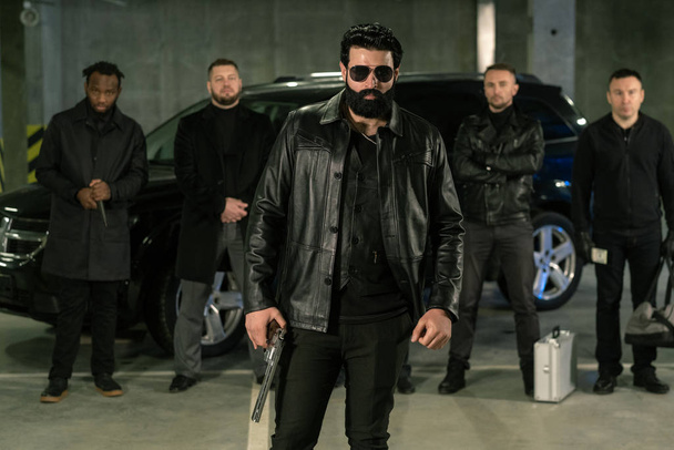 Bearded criminal authority in sunglasses, black leather jacket and jeans holding handgun with his gang on background - Photo, Image