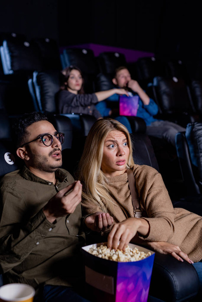Tense young couple with box of popcorn watching action movie while sitting in dark cinema in front of screen - Valokuva, kuva