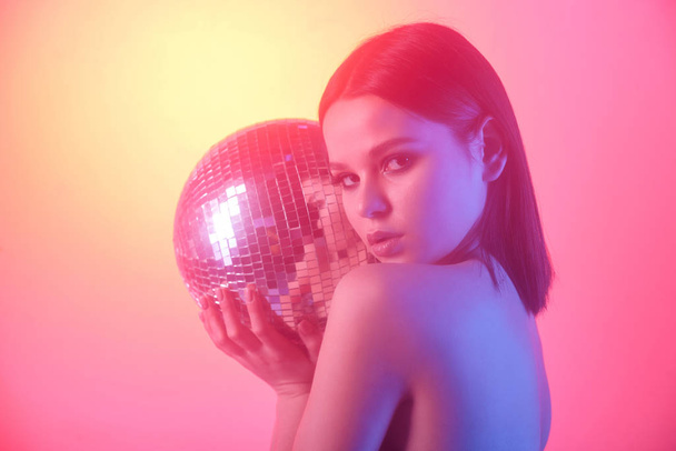 Portret van serieus naakt meisje houden disco spiegel bal tegen licht geel en roze achtergrond - Foto, afbeelding