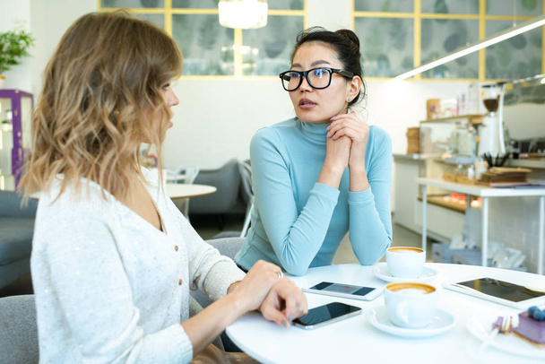 Twee jonge interculturele zakenvrouwen in casualwear die discussiëren over werkplekken of plannen terwijl ze koffie drinken aan tafel in café - Foto, afbeelding
