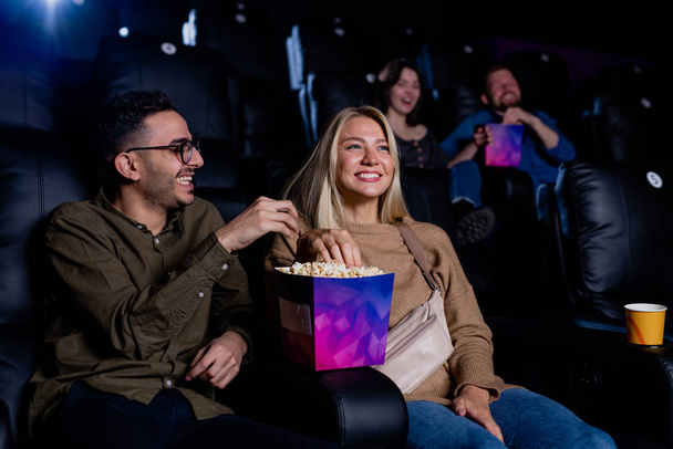 Веселая молодая пара ест кукурузу, сидя в кино во время фильма и наслаждаясь отдыхом - Фото, изображение