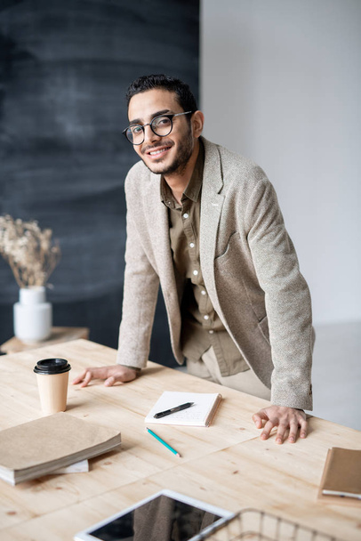Fiatal mosolygós vállalkozó vagy menedzser a formalwear néz rád, miközben hajol a fa asztal munka közben - Fotó, kép