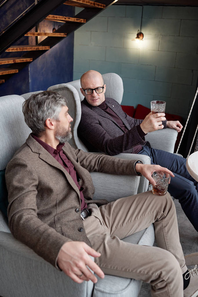 Elgondolkodtató, stílusos középkorú férfiak kényelmes karosszékben ülve és kemény alkoholt iszogatva, miközben beszélgetnek - Fotó, kép