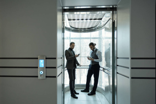Zwei junge gut gekleidete männliche Unternehmer mit mobilen Geräten im Aufzug eines modernen Geschäftszentrums - Foto, Bild