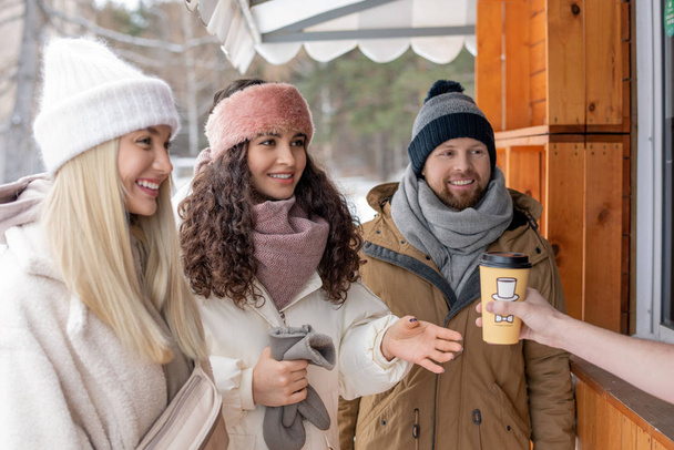 Ευτυχισμένοι νέοι φίλοι σε casual χειμωνιάτικα ρούχα αγοράζουν ζεστό καφέ σε ξύλινο πάγκο την ημέρα του χειμώνα, ενώ ψύξη έξω - Φωτογραφία, εικόνα