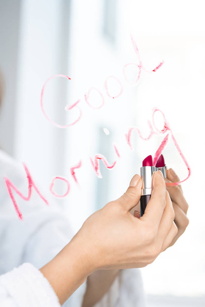 Mano de mujer joven sosteniendo lápiz labial de color carmesí cerca del espejo mientras escribe buenos días
 - Foto, imagen