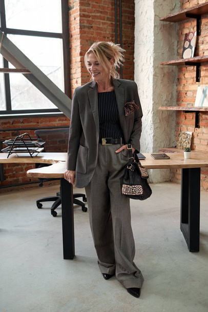 Счастливая владелица бизнес-компании, стоящая за столом и держащая стильную сумку в лофт-офисе
 - Фото, изображение