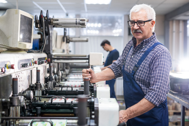 Σοβαρός ηλικιωμένος άνδρας με άσπρα μαλλιά στη συνολική λειτουργία εκτυπωτικής μηχανής στο εργοστάσιο - Φωτογραφία, εικόνα