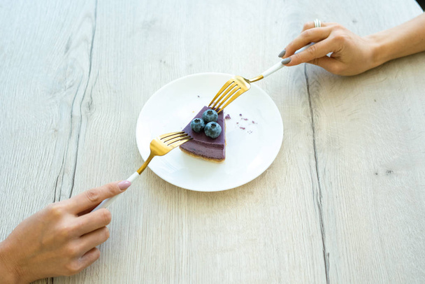 Hands of two young females with forks taking piece of appetizing blueberry dessert while sitting by wooden table - Photo, Image