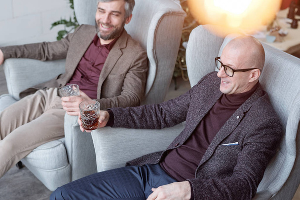 Nad pohledem veselých podnikatelů středního věku ve stylových bundách sedících v křeslech a popíjejících whisky - Fotografie, Obrázek