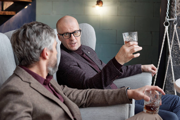 Moderní podnikatelé středního věku sedí v křeslech v restauraci a diskutují o obchodu se sklenicí whisky - Fotografie, Obrázek