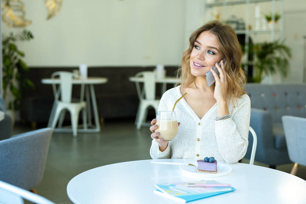 Gelukkig meisje met een glas cappuccino en bosbessen dessert praten met de smartphone terwijl ontspannen in cafe - Foto, afbeelding