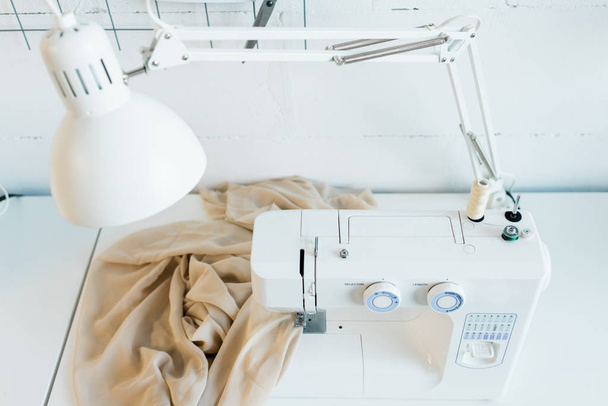 Вид зверху на робоче місце з електричною швейною машиною, настільною лампою та тканиною
 - Фото, зображення
