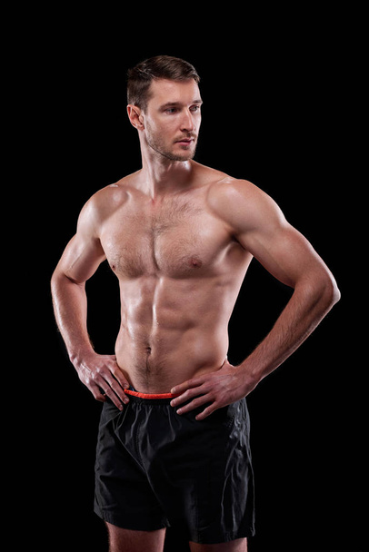 Young muscular shirtless male athlete keeping his hands on waist while standing in front of camera in isolation - Фото, изображение