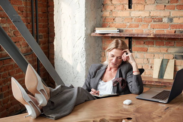 Inhoud moderne volwassen zakenvrouw houdt voeten op tafel in kantoor en het controleren van notities in planner  - Foto, afbeelding