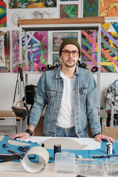 Porträt eines ernsthaften jungen kreativen Lederarbeiters mit Brille und Hut, der am Schreibtisch mit Kartonmustern steht - Foto, Bild