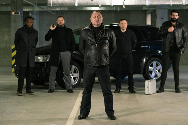 Reifer Mischlingsgangster oder Krimineller in schwarzer Lederjacke und Jeans steht vor seiner Bande auf dem Parkplatz - Foto, Bild